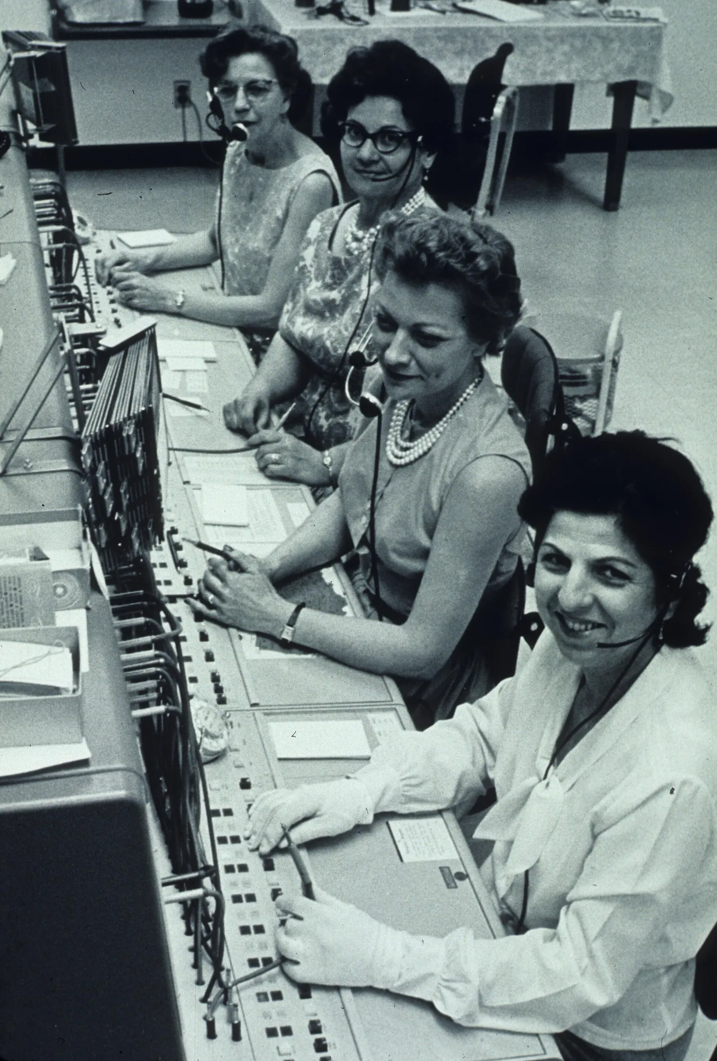 Four switchboard operators wearing headsets at a long cord switchboard, smiling at the camera
