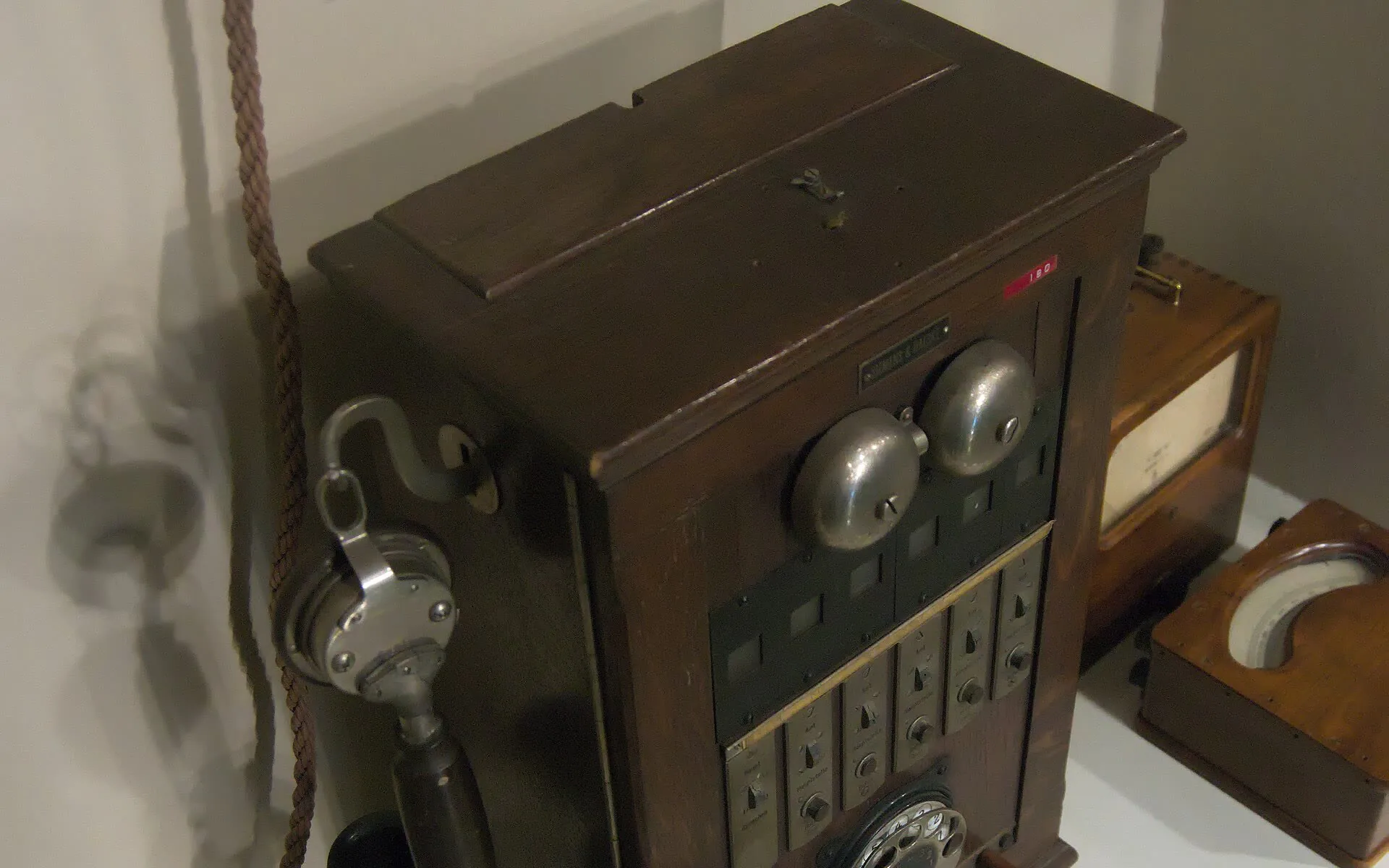 A wooden Siemens and Halske telephone switchboard with two bells, a handset and cord jacks, on a museum shelf